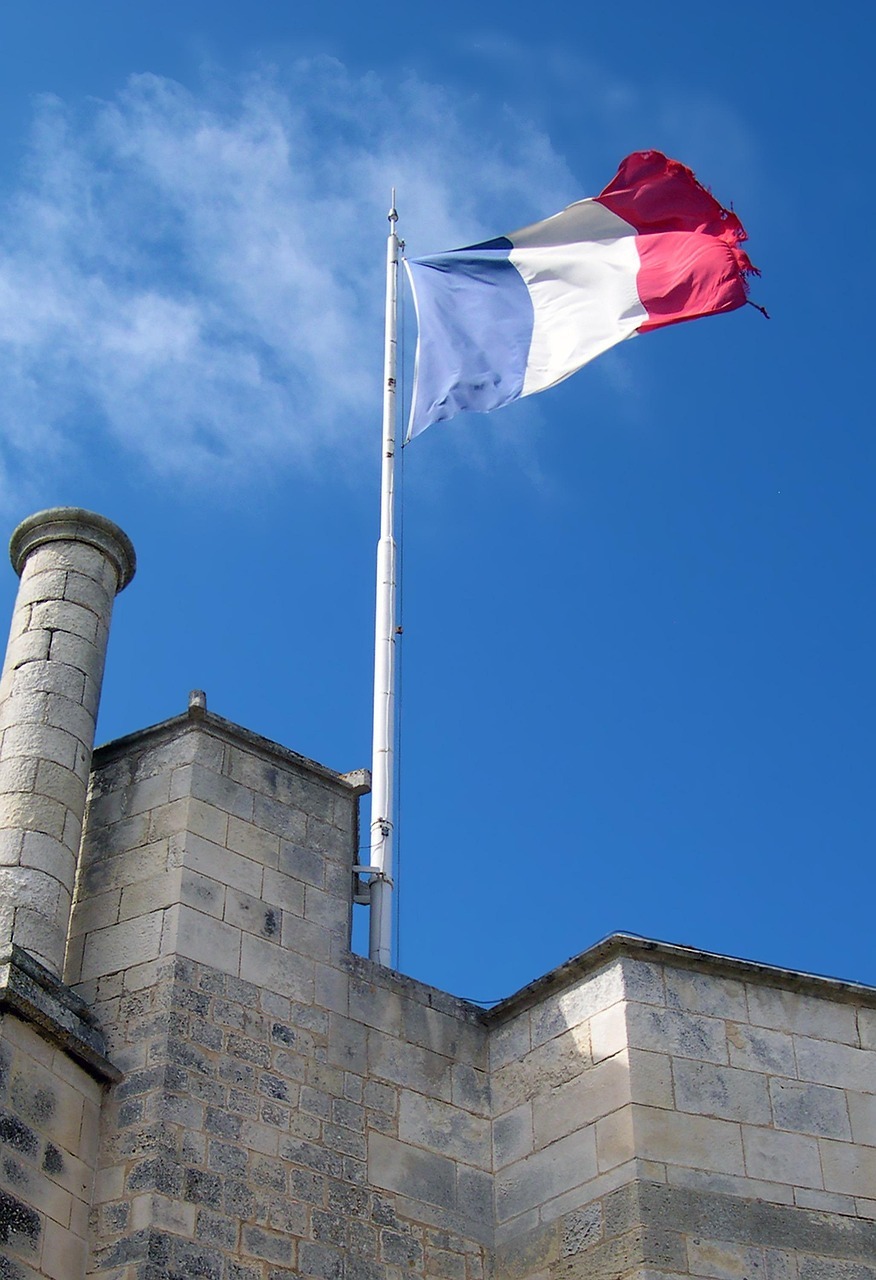 Drapeau français aux couleurs bleu, blanc et rouge, flottant au sommet d'un grand mât sur un bâtiment historique en pierre.