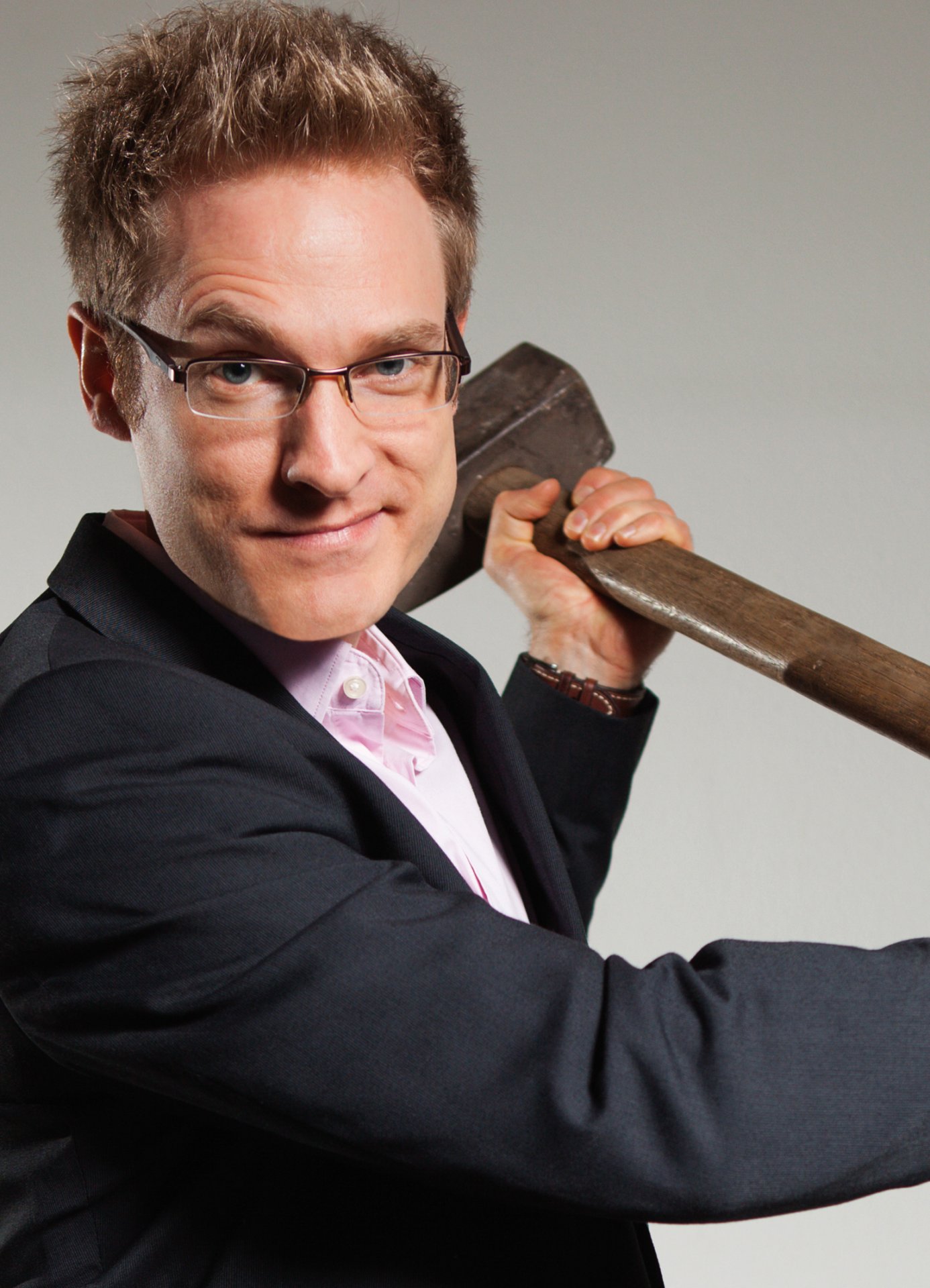 Personne aux cheveux blonds et courts portant un costume noir et une chemise rose clair, tenant une lourde hache sur l'épaule.