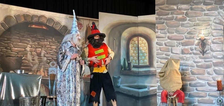 Scène de théâtre avec trois personnages en costume : un magicien avec une longue barbe blanche et un chapeau pointu, un personnage avec une chemise rouge, un chapeau noir et une barbe, et un personnage assis avec un sac sur la tête et un pantalon rouge.