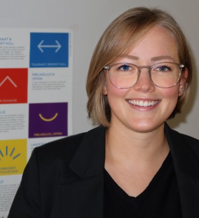 Mme Hannig porte un blazer noir, une chemise noire et des lunettes et se tient devant un mur avec une affiche colorée.