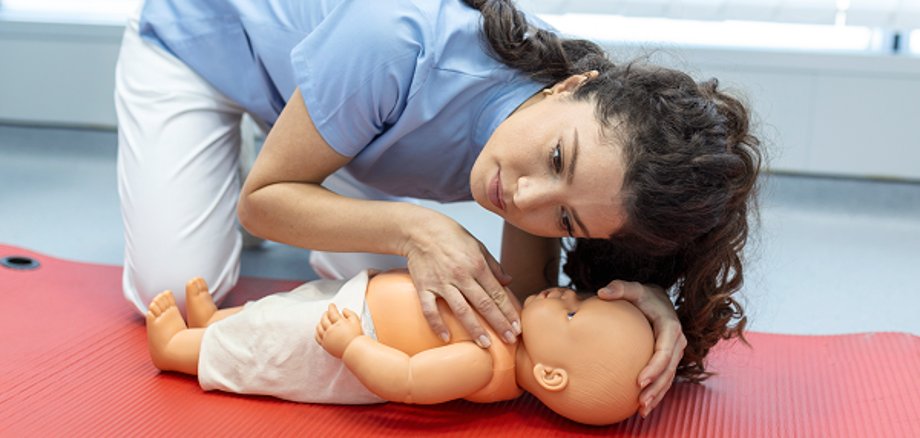Une personne pratique le bouche-à-bouche sur un bébé mannequin posé sur un tapis rouge dans le cadre d'un cours de secourisme