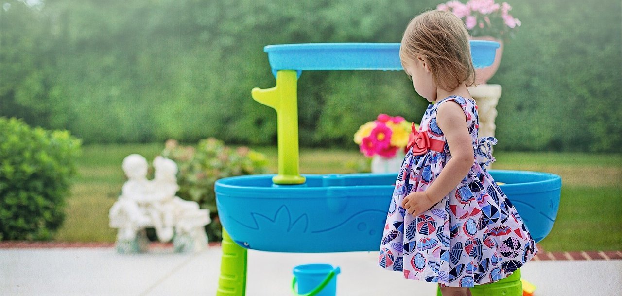 Une petite fille en robe à motifs et sandales se tient devant une table de jeu bleue et verte avec un seau dans le jardin.