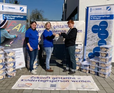 Trois personnes, devant des banderoles de la ville de Rodgau, fournissent des informations sur l'accueil des enfants en crèche, avec à côté d'elles des piles de cartons remplis de jouets