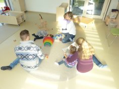 Trois adultes et trois enfants assis sur le sol d'une salle de groupe lumineuse d'une crèche, jouant et lisant ensemble