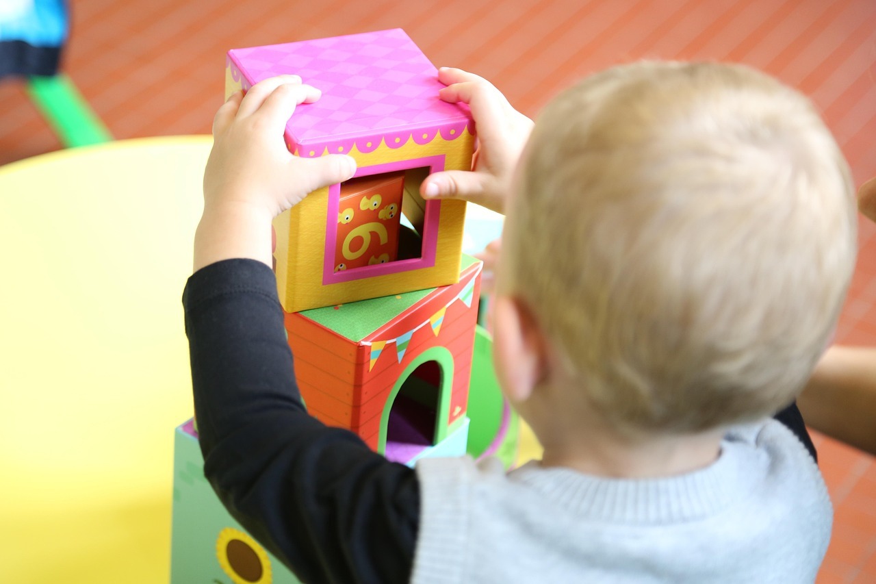 Un jeune enfant aux cheveux blonds empile des cubes de jeu colorés sur une table. Les cubes sont de couleurs et de motifs différents.