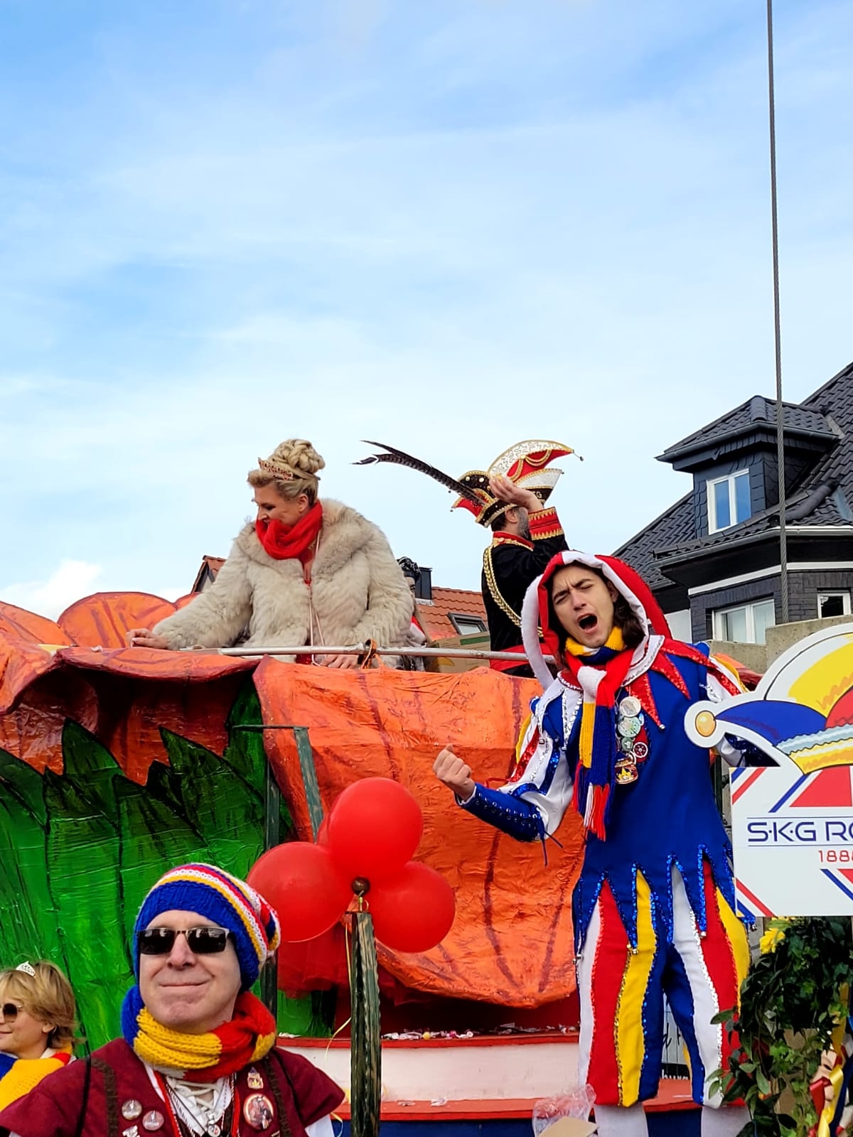 Couple princier du carnaval en costumes colorés, coiffé d'un bonnet de fou et portant une plume, sur un char entouré de ballons rouges et de spectateurs.