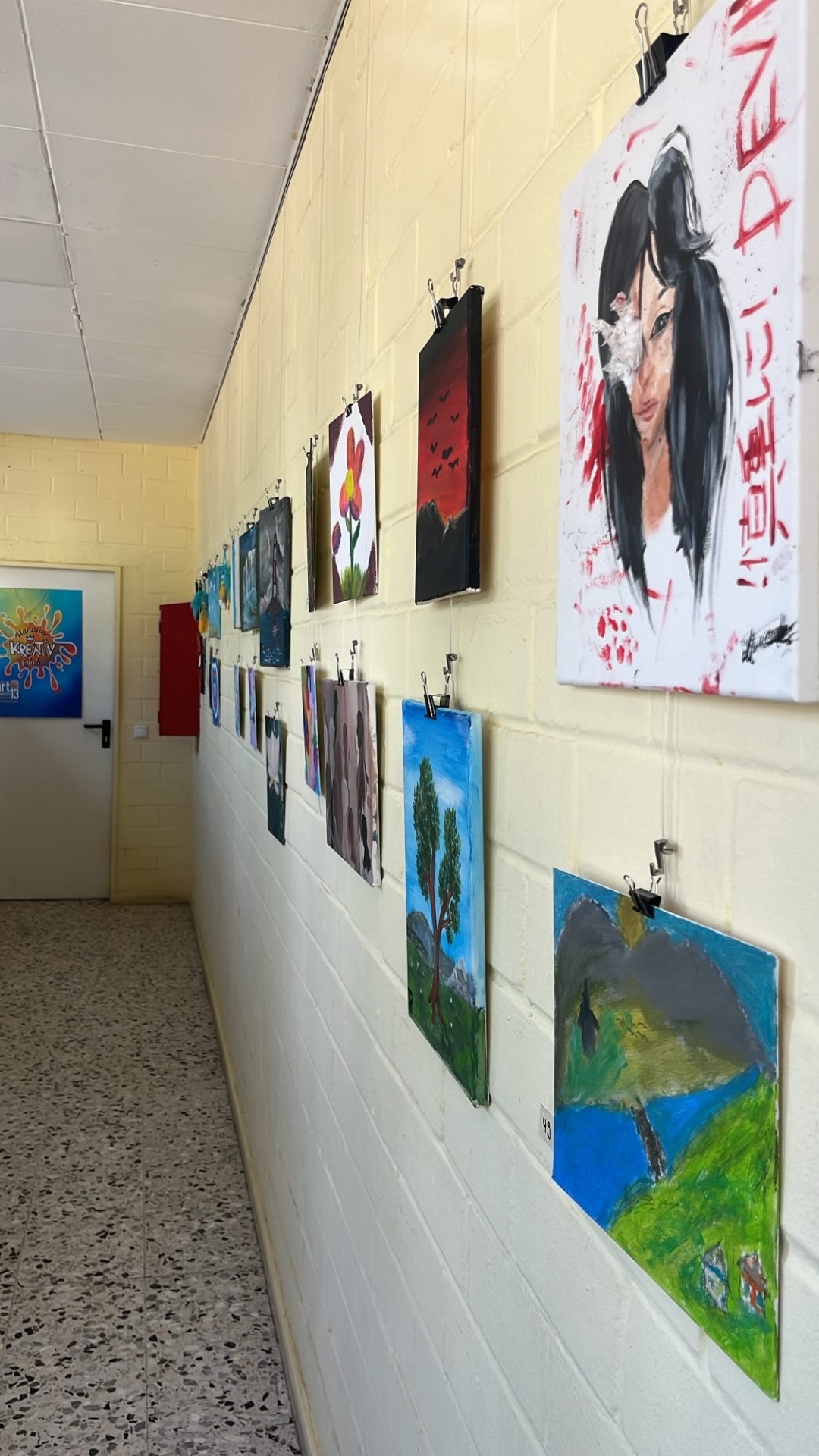 Des œuvres d'art de la JugendArt sont accrochées sur un mur blanc dans un couloir, dont un portrait de femme aux cheveux noirs sur fond rouge.