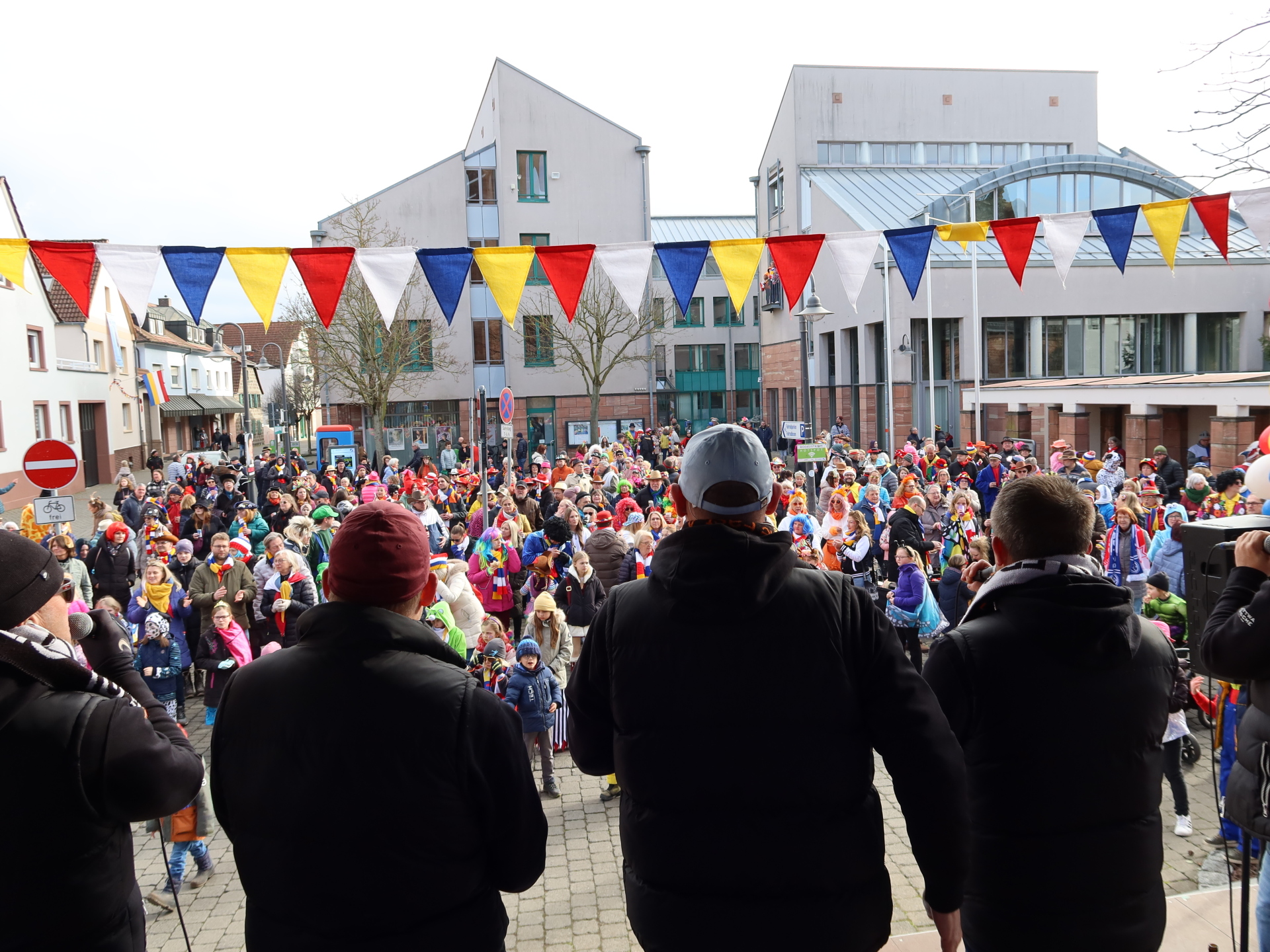 Foule devant l'hôtel de ville lors du carnaval, des fanions colorés surplombent la scène. Au premier plan, trois personnes portent des vestes noires, dos à la caméra.