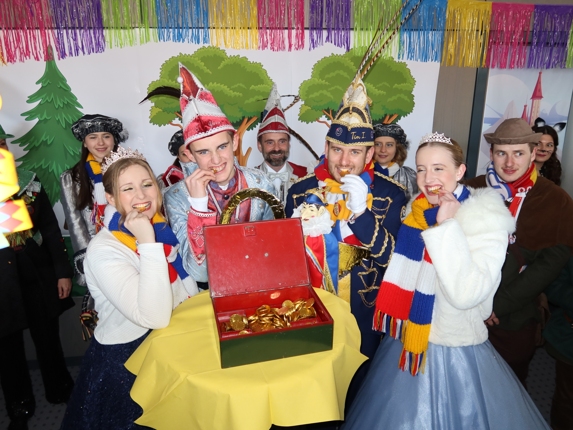 Six personnes en habits de fête, dont un prince et une princesse, se tiennent autour d'une table ronde avec une boîte verte. En arrière-plan, on peut voir des décorations colorées et des silhouettes de sapins de Noël.