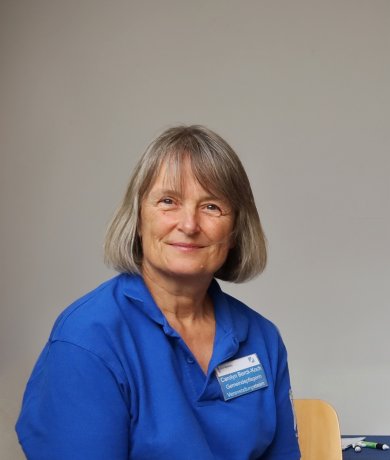Portrait de l'administratrice municipale Berck-Koch devant la salle de réunion du conseil municipal. Elle a des cheveux gris blonds et porte un chemisier bleu.