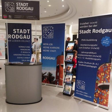 Présentoir bleu de la ville de Rodgau avec une publicité pour les métiers de l'éducation. Des brochures sont disposées à côté. D'autres exposants sont visibles à l'arrière-plan.
