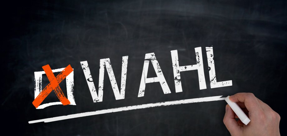 Main écrit à la craie blanche sur un tableau noir le mot 'ÉLECTION' précédé d'une croix rouge dans une case.