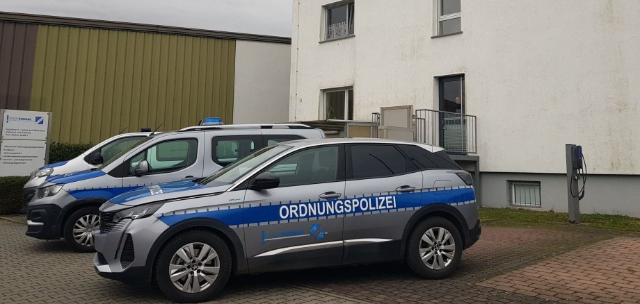 Trois véhicules de la police de l'ordre public sont garés devant un bâtiment blanc à plusieurs étages et au toit gris, par une journée nuageuse.