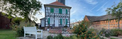 Rektor-Geißler-Haus avec jardin Rektor-Geißler-Haus à Dudenhofen, une maison historique à colombages avec des volets verts et blancs et une tour, entourée d'un jardin avec des fleurs et un banc blanc.