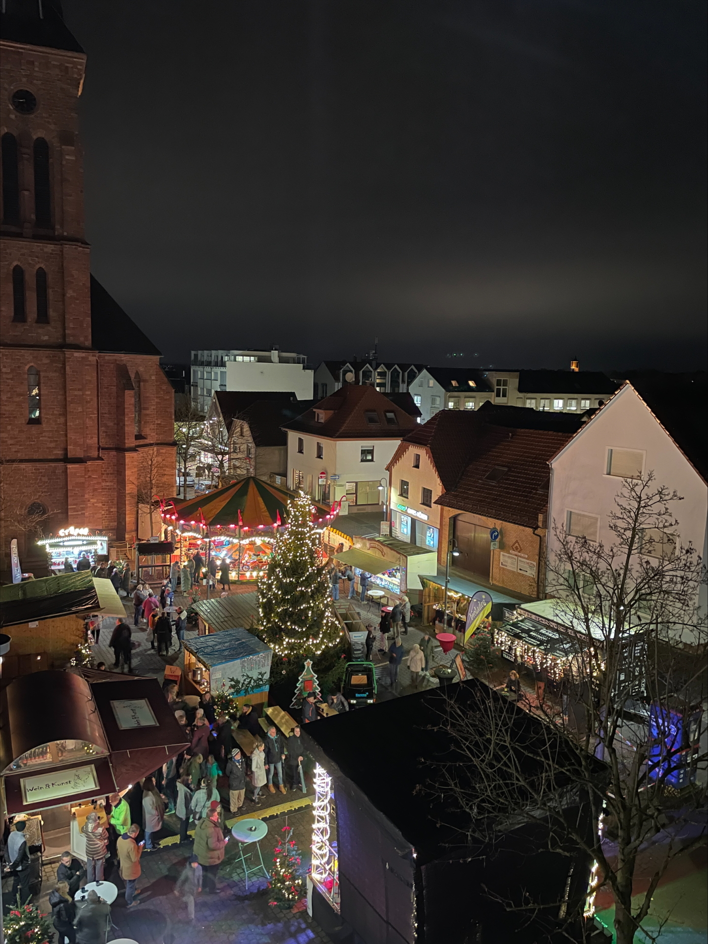 Marché de Noël à Jügesheim Marché de l'Avent en soirée sur la place de la mairie de Rodgau, avec un sapin de Noël illuminé, un manège et des stands de marché devant des bâtiments historiques.