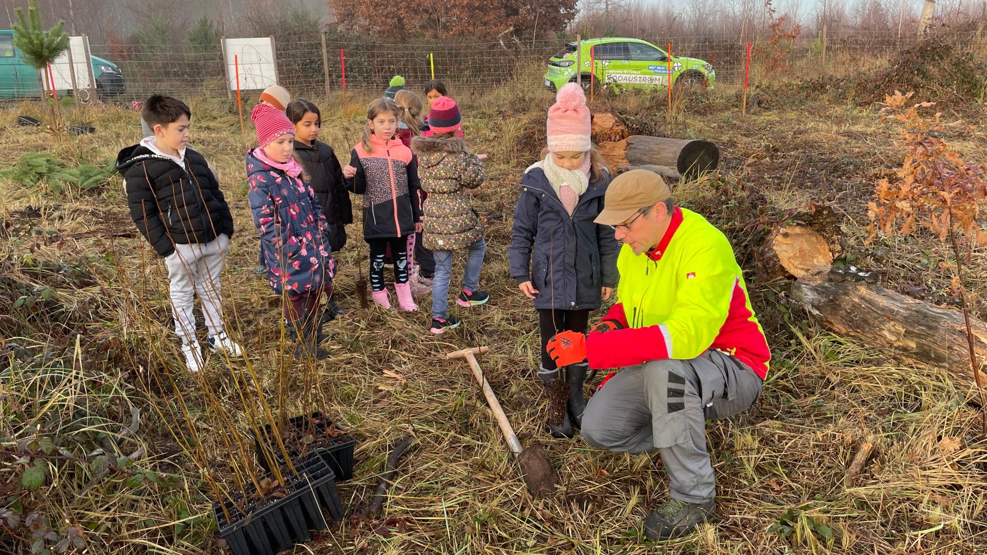 Gruppo di bambini e un dipendente comunale in tenuta di avvertimento piantano giovani alberi in un campo con attrezzi e piantine.