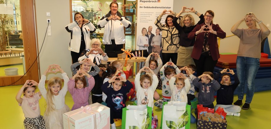 Gruppo di adulti e bambini al Kita 18, che formano cuori con le mani, davanti a diversi pacchi regalo e a uno striscione Piepenbrock.