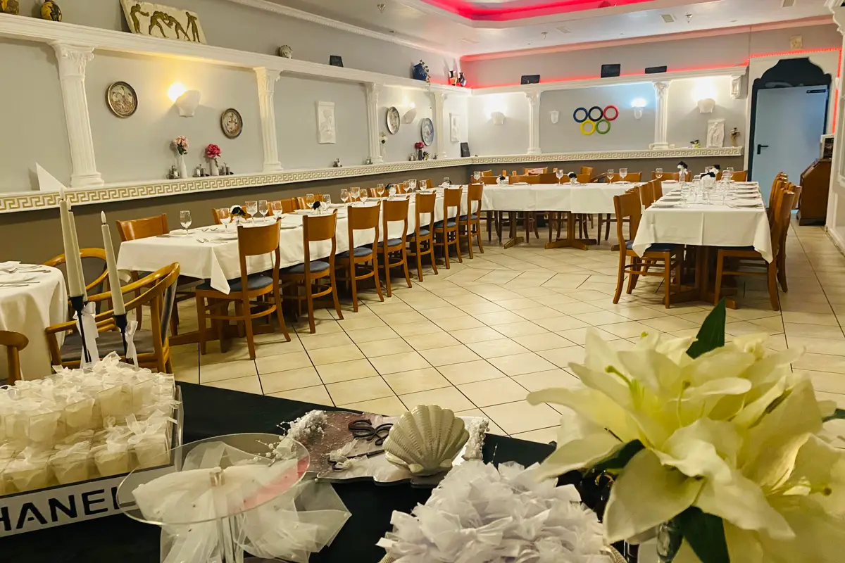 Ampia sala da pranzo decorata a festa con diversi tavoli lunghi, pavimento in legno e tovaglie bianche, decorata con fiori e orologi da parete.