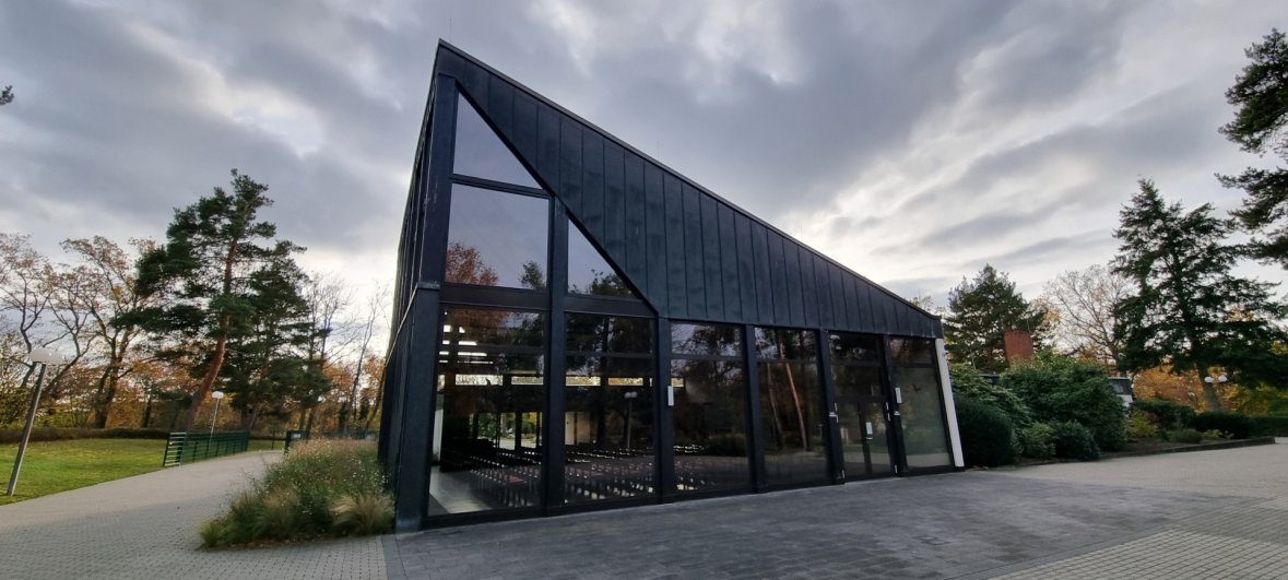 Sala cimiteriale del cimitero forestale di Jügesheim Edificio moderno e affusolato della sala cimiteriale con tetto nero e grandi facciate in vetro, circondato dagli alberi e dai camminamenti del cimitero forestale.