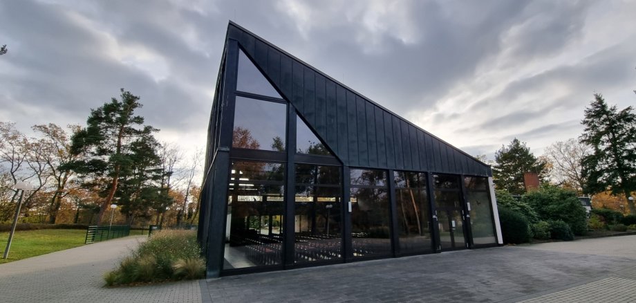 Edificio moderno e affusolato della sala cimiteriale con tetto nero e grandi facciate in vetro, circondato dagli alberi e dai camminamenti del cimitero forestale.