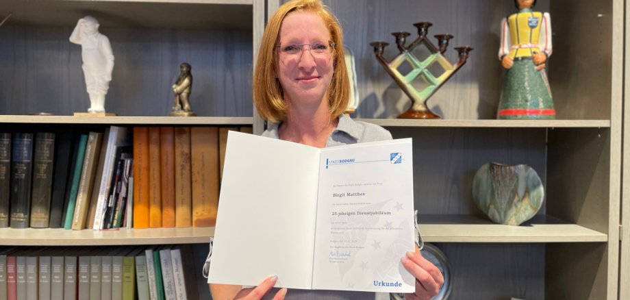 Birgit Mattes tiene in mano un certificato con il testo "25 anni di servizio". Sullo sfondo, libri, una statua e una vetrina decorativa.