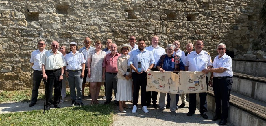 Un gruppo di 14 persone si trova davanti a un muro di pietra. Alcune tengono in mano un grande foglio di carta piegato con dei disegni. L'abbigliamento varia da formale a casual.