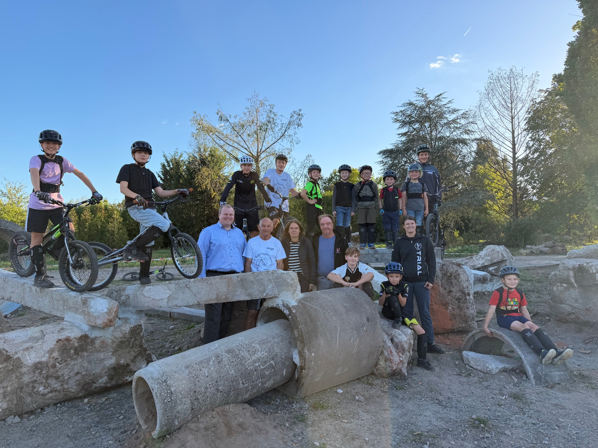 Gruppo di bambini e adulti con caschi e biciclette su e intorno a grandi tubi di cemento e rocce all’aperto