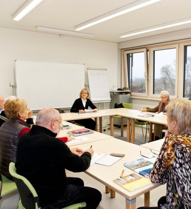 Un gruppo di anziani è seduto a tavoli disposti a forma di U in un’aula luminosa con grandi finestre e scrive o ascolta