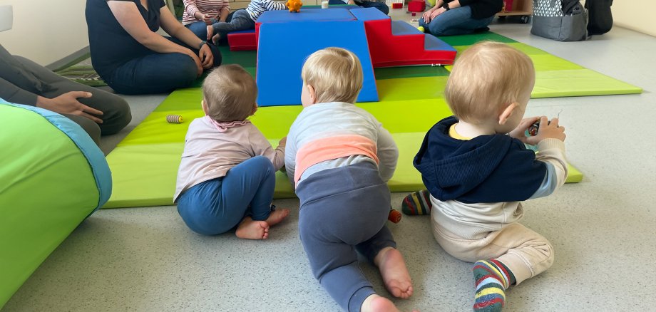 Tre bambini Tre bambini piccoli sono seduti e gattonano su un tappeto verde chiaro. Sullo sfondo si vedono altri bambini e adulti seduti attorno a una struttura da gioco blu e rossa. I bambini indossano abiti di vari colori, tra cui calzini e maglioni a righe. Sul tappeto sono sparsi dei giocattoli.
