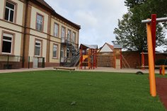 Parco giochi con struttura per arrampicata e scivolo accanto a un edificio scolastico a due piani con scala di fuga esterna
