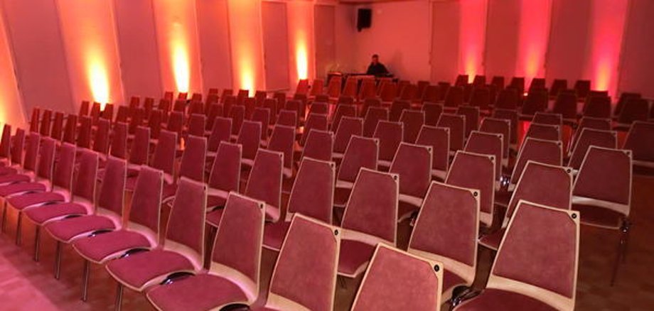 Centro comunitario di Niederroden Sala del centro comunitario di Niederroden con diverse file di sedie rosse e illuminazione a parete rosso-arancio