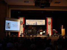 Palco con pianoforte, batteria e microfoni alla cerimonia di premiazione del Premio di promozione culturale della città di Rodgau, schermo a sinistra con discorso di benvenuto di Lars Neumann
