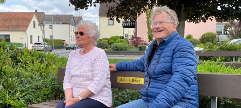 Babbelbank a Hainhausen Una signora e un signore sono seduti su una panchina in una piazza di Hainhausen ricca di verde.