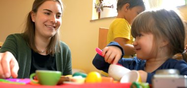 PivA L'insegnante (a sinistra) gioca con due bambini (a destra) in una cucina da gioco. Sullo sfondo si vede l'interno di un asilo nido.