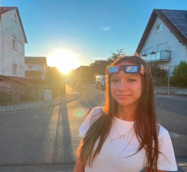 Anna Lena Hitzel Una ragazzina con i capelli lunghi e una maglietta bianca ha gli occhiali per l’eclissi solare sulla fronte ed è in piedi su una strada al tramonto