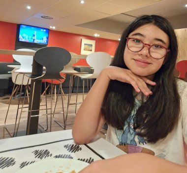 Asuman Yildiz Una ragazzina con lunghi capelli scuri e occhiali rossi è seduta a un tavolo in un bar e tiene il mento appoggiato sulla mano