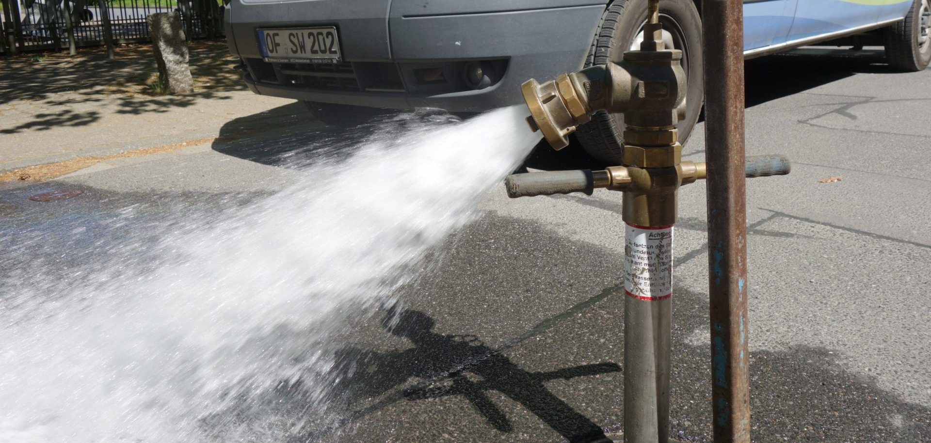 Strumień wody tryska z hydrantu na chodnik. W tle widać białą furgonetkę z zielonymi i czerwonymi oznaczeniami.