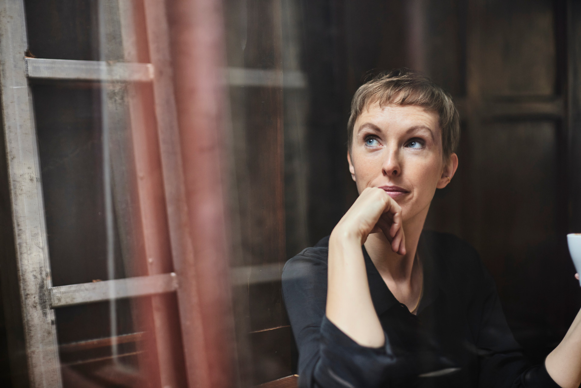 Osoba o krótkich blond włosach siedzi w ciemnym pokoju. Ma na sobie czarną bluzkę i trzyma kubek w prawej ręce. W tle widać drewnianą drabinę i ścianę.