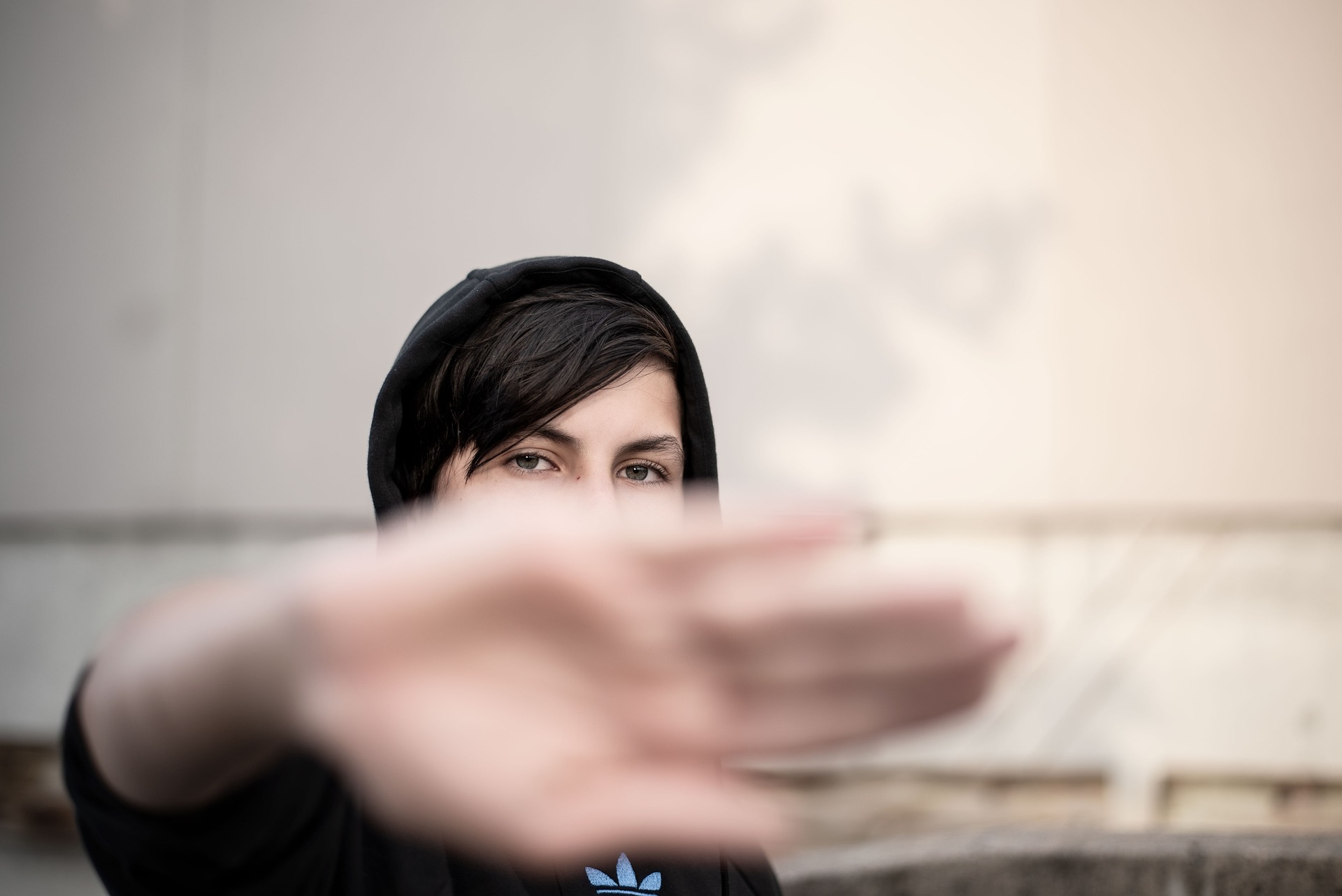 Kobieta w czarnej kurtce z kapturem trzyma dłoń do kamery, rozmyte tło ze ścianą i graffiti