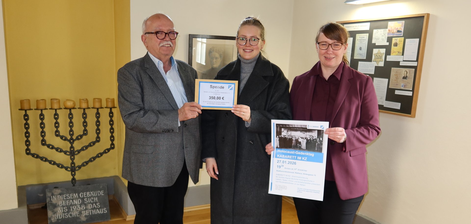 Trzy osoby stoją w pokoju z menorą i tablicą pamiątkową, trzymając tabliczkę z napisem "Darowizna 350,00 €" i plakat wydarzenia upamiętniającego Holokaust.