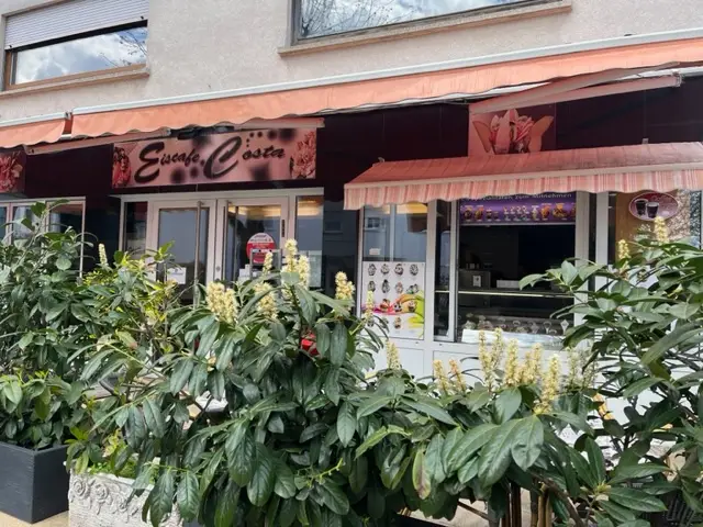 Lodziarnia z napisem "Eiscafe Costa" za zielonymi roślinami w donicach