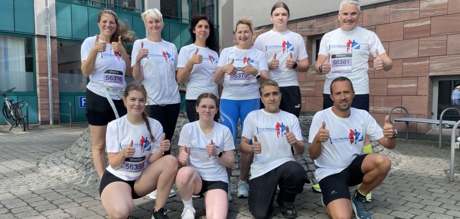 Uczestnicy biegu organizowanego przez miasto w ramach imprezy JP Morgan Chase Run Grupa dziewięciu pracowników miejskich w białych koszulkach z logo JP Morgan Chase pozuje z uniesionymi kciukami na brukowanym dziedzińcu ratusza