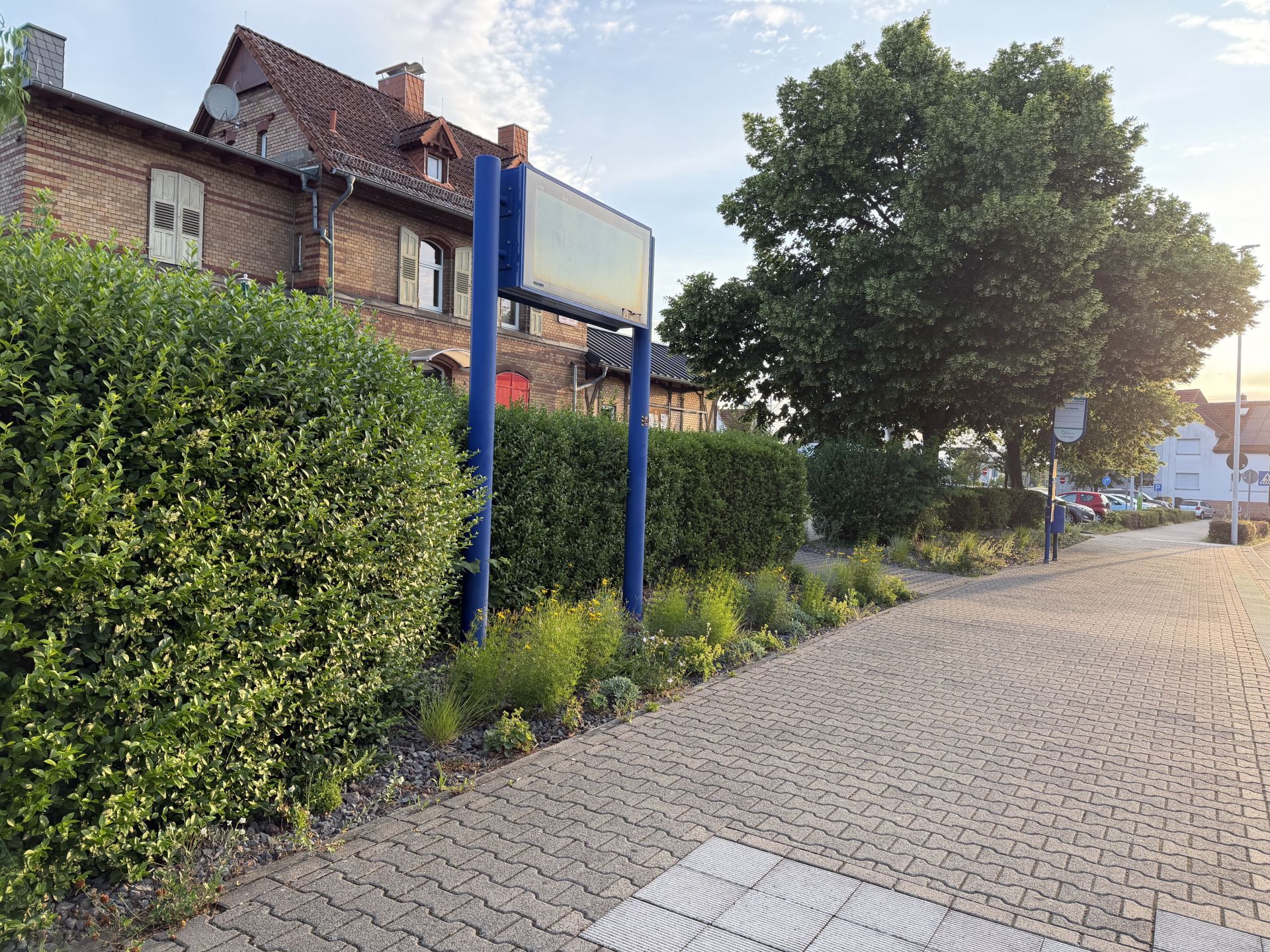 Widok na stację kolejową w Nieder-Roden Widok na stację Nieder-Roden z niebieskim znakiem przed żywopłotem, brukowanym, czystym chodnikiem i dużym drzewem w tle