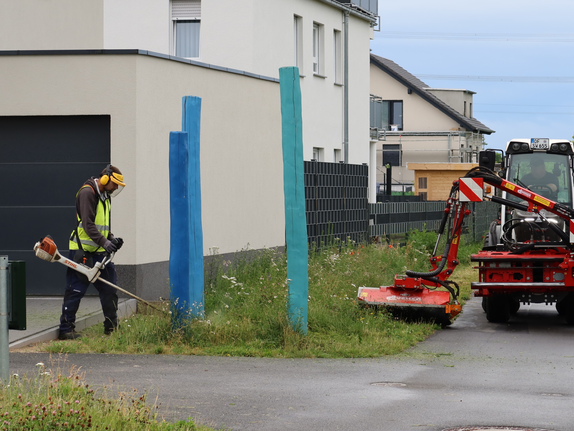 Dział zieleni w Zakładach Komunalnych w Rodgau Pracownik działu zieleni przedsiębiorstwa Stadtwerke Rodgau kosi kosiarką spalinową trawnik przy budynku mieszkalnym, tuż obok dwóch niebieskich drewnianych słupków; po prawej stronie na chodniku i ścieżce rowerowej stoi kosiarka zamontowana na ciągniku.
