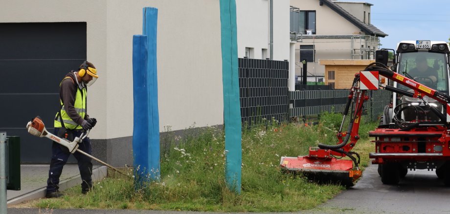 Dział zieleni w Zakładach Komunalnych w Rodgau Pracownik działu zieleni przedsiębiorstwa Stadtwerke Rodgau kosi kosiarką spalinową trawnik przy budynku mieszkalnym, tuż obok dwóch niebieskich drewnianych słupków; po prawej stronie na chodniku i ścieżce rowerowej stoi kosiarka zamontowana na ciągniku.