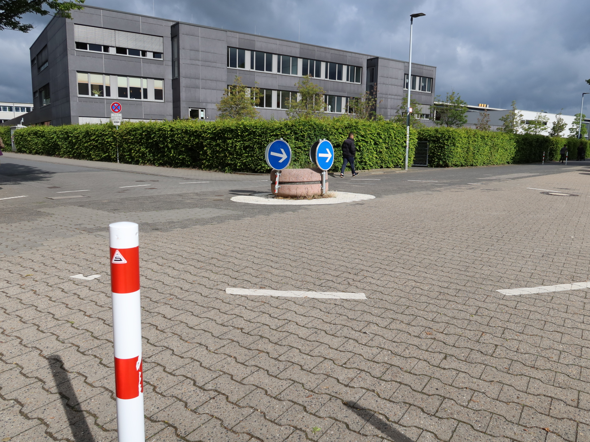 Małe rondo z dwiema niebieskimi strzałkami kierunkowymi na platformie z okrągłą wyspą centralną między Kita 18 a szkołą Georga Büchnera, otoczone utwardzonym terenem i wielopiętrowym budynkiem szkoły w tle.