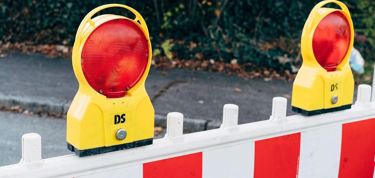 Utrudnienia w ruchu Dwa żółte światła ostrzegawcze z czerwonym światłem na białej barierze z czerwonymi i białymi paskami. W tle widoczne są zielone rośliny.