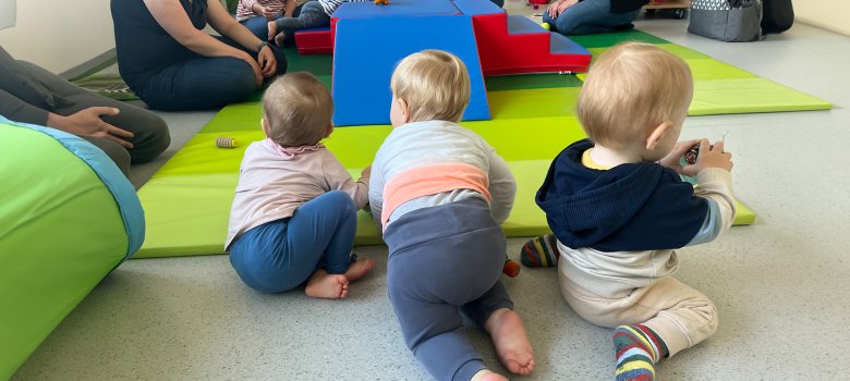 Trójka małych dzieci siedzi i raczkuje na jasnozielonej macie. W tle widać kolejne dzieci i dorosłych siedzących wokół niebiesko-czerwonej konstrukcji do zabawy. Dzieci mają na sobie różnokolorowe ubrania, w tym skarpetki w paski i swetry. Na macie leżą rozrzucone zabawki.
