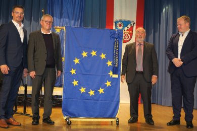 Burmistrz Breitenbach, radny Schüssler i radny Kaiser stoją na scenie; w centrum znajduje się flaga europejska