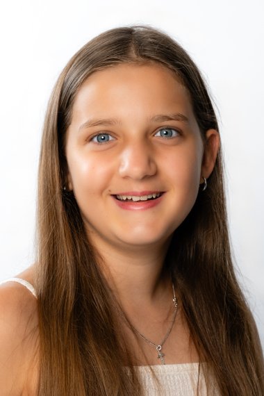 Portret uśmiechniętej dziewczyny o długich brązowych włosach, ubranej w białą bluzkę i naszyjnik z zawieszką w kształcie krzyża